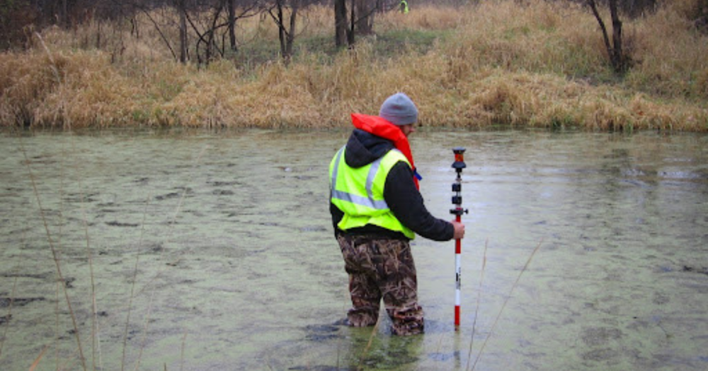 A man standing in a pond holding a prism for a total station survey. A man standing in a pond holding a prism for a total station survey.